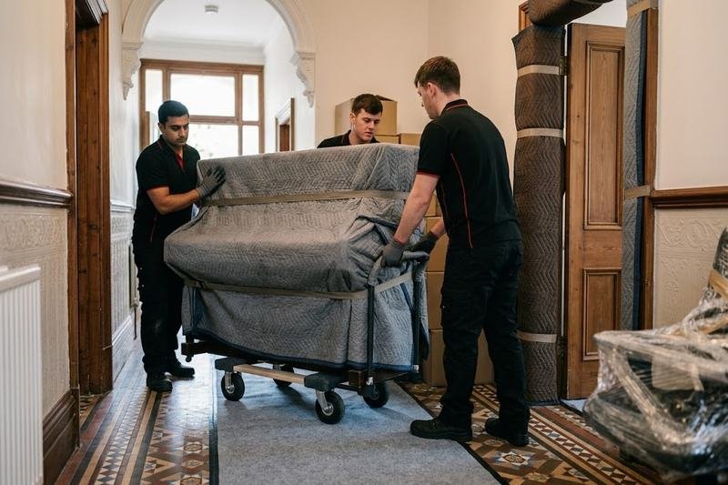 Three movers wheeling a blanket-wrapped upright piano on a dolly through a Victorian hallway